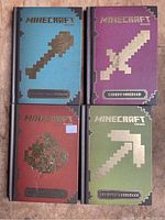 Photo of all four Minecraft handbooks showing distinct colored covers with pixel art tools: shovel (blue), sword (purple), redstone (red), pickaxe (green).