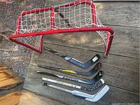 Mini hockey sticks and mini hockey net placed outdoors on a wooden surface and gravel ground, showing 5 sticks and a red framed net with white netting.