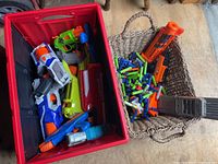 Top-down photo showing several Nerf blasters inside a red storage bin and a pile of Nerf darts and an orange dart ammo clip in a woven basket to the right.