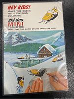 Front of vintage 1970 Ski-Doo Mini Transfers booklet with colorful winter adventure scene, a cabin by a pond, skiers, and snowmobiles; includes text inviting kids to create scenes with transfers.