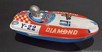 Side view of vintage tin boat toy showing red, white, and blue colors, with 'DIAMOND' and 'Y-22' markings and attached metal driver figure wearing a shiny silver helmet.