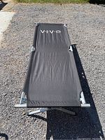 Top view of the VIV branded black fabric stretched on the metal frame of camping cot on gravel ground.