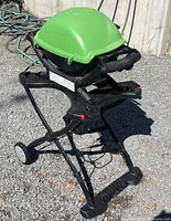 Overall view of the green Weber Q 1200 portable BBQ grill mounted on a black metal foldable stand with wheels. Shows the grill’s compact design and stand construction.