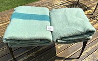 Two folded green wool blankets with teal stripes, stacked on a metal chair outdoors.