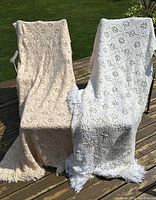Both crocheted blankets draped on chairs outdoors showing size, fringe on 3 sides and design.