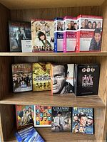 Photo of nine DVD boxed sets on wooden shelves showing variety of genres including comedy, mystery, western, and classic movie collections, along with an Allsop DVD Lens Cleaner.