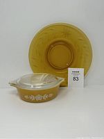 Pyrex lidded casserole dish in Butterfly Gold pattern alongside a goldenrod colored depression glass plate with auction lot number card.