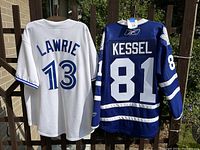 Front and back view of Toronto Blue Jays white jersey and Toronto Maple Leafs blue jersey hanging on railing outside.
