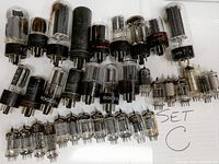 Various vacuum tubes of different sizes arranged on a white surface, showing clear glass envelopes and metal pins, marked with 'SET C' on a paper.