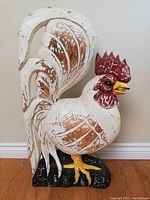 Front view of large carved wooden rooster painted white with brown feather detailing, red comb, yellow beak and feet, standing on black base.
