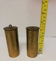 Two brass cylindrical clock weights standing upright next to a measuring tape showing height approximately 4.5 inches.
