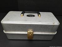 Closed view of vintage $ fishing lure box showing textured silver metal surface, black plastic handle, and lock latch.