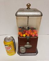 Front view of Cadillac Jr. small universal vending gumball machine, showing metal top with key, transparent container with colorful gum balls, coin slot and handle, metal base, and a Canada Dry soda can for size reference.