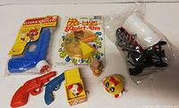 Full view of all toys in lot including three NOS items in original packaging: Crazy Gun water pistol, Mr. Pac-Man Squid Oyn card game, and several small toy pistols, character toys, and wind-up toys.