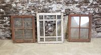 Front view of three antique wooden sash windows against a stone wall and wood floor: two brown 2-over-2 barn windows flanking one white multi-pane house window.