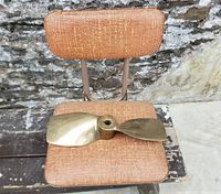 Front view of brass boat propeller resting on the seat of the collapsible chair with textured orange upholstery and metal frame.
