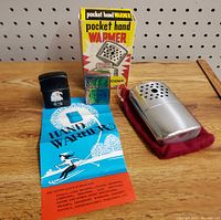 Lot overview photo displaying two Harley-Davidson lighters, the pocket hand warmer with original box, red pouch, and instruction sheet on a wooden surface against pegboard background.