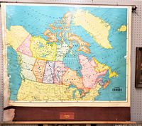Front view of large school map of Canada with pastel colored provinces, a dark brown fabric edging at bottom with a red label for publisher and original metal 'Canada' tag on mechanism.