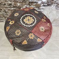Top view of the round leather ottoman showing patchwork of brown and red leather panels with embroidered flower patterns and dotted accents.