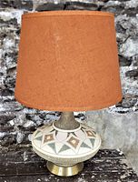 Front view of mid-century ceramic flying saucer styled table lamp with burgundy burlap shade against stone background on wooden surface.