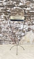 Mid-century modern bird cage mounted on a thin black metal hooped stand in front of a stone wall. Cage is rectangular with wire bars and a beige plastic base with decorative corner accents. Plastic feeders are affixed inside the cage.