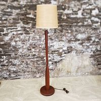 Full view of the mid-century teak floor lamp with burlap shade against a textured stone wall background, showing the lamp standing upright on the floor.