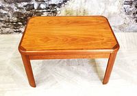 Frontal view of the mid-century teak small coffee or side table showing the rectangular top with rounded edges and wide rounded legs, in front of a textured stone wall background.
