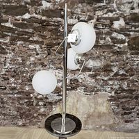 Front view of the lamp showing three white glass nipple shades mounted on a chrome pole with circular chrome base, against a stone wall background.