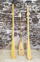 Front view of the pair of long yellow lacquered wooden boat oars alongside the shorter wooden branded paddle, all standing upright against a stone wall.