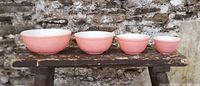 Front view of four graduated pink Pyrex bowls displayed in line from largest to smallest on a rustic wooden bench against a stone wall background.