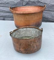 Two antique hammered copper pans or buckets placed on a white surface against a stone background. Larger smooth copper bucket at the back; smaller hammered copper pan with tin lining in front.