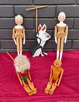 Front view of five wooden marionette puppets on red cloth with brick background: two human figures, one rabbit figure with control bar, and two wooden dolls, one with fur head covering.