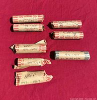 Seven cylindrical rolls of pennies, wrapped in paper, each labeled with country and date range, arranged on a red fabric background.