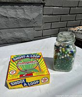 Photo showing the jar of marbles next to the Shoot-A-Loop marble game box outdoors against a stone wall background.