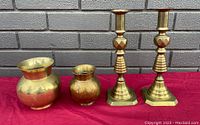 Photo showing all four items: two brass candlesticks and two brass vases of different sizes against a brick wall on red cloth.