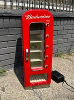 Front and full view of the bright red Budweiser mini fridge with glass door, shelves and dispenser style knobs on the side, showing the budweiser logo and ice cold text on bottom.