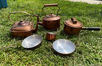 Photo showing entire set of copper kitchenware on grass including kettles, saucepan, small pot, and frying pans.