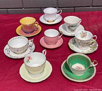 Seven vintage fine bone china tea cups and matching saucers displayed on red cloth against brick background, various colors and patterns visible.