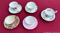 Five tea cups and saucers arranged on red fabric background showing varied floral patterns and gold accents.