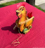 Front and side view of the yellow wooden cackling hen pull toy sitting on red cloth outdoors, showing string attached and visible wheels.