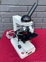 Front angle view of white Leitz HM-Lux microscope on red cloth, showing body, stage, objective lenses, and base with power cord.