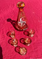 Full set photo showing orange glass conical decanter with matching five shot glasses on red background, all featuring white printed festival logo.