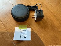 Black Amazon Echo Dot with four buttons on top next to a black power adapter and auction lot number 112 card on a wooden table.