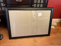 Photo showing the black rectangular picture frame standing on the floor against a wooden furniture and red wall background.