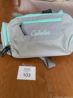 Front view of Cabela's gray and teal fishing tackle bag showing front zippered pocket with logo and shoulder strap.