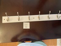 Photo showing a white wooden coat rack with six silver metal hooks mounted horizontally on a dark surface with a small numbered card beneath it.