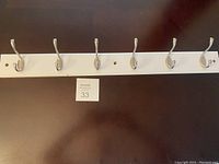 Photo of a white wooden coat rack with 6 silver hooks laid on a dark surface, showing the entire length and detail of hooks.