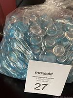 Clear plastic bag filled with numerous light blue glass stones, glossy and translucent. Stones appear smooth and rounded, suitable for decoration.