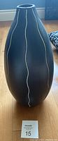 Tall safari style decorative vase with black matte finish and vertical white wavy lines standing on wooden floor near window