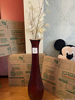 Tall red glass vase with white acrylic leaf branches displayed upright in room with moving boxes and Mickey Mouse plush in background